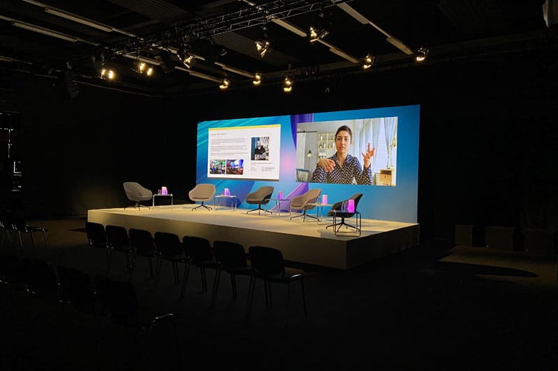 Bühne mit 5 Sesseln und einer LED Wand auf der links eine Powerpoint Präsentation und rechts die Referierende Person eingeblendet ist